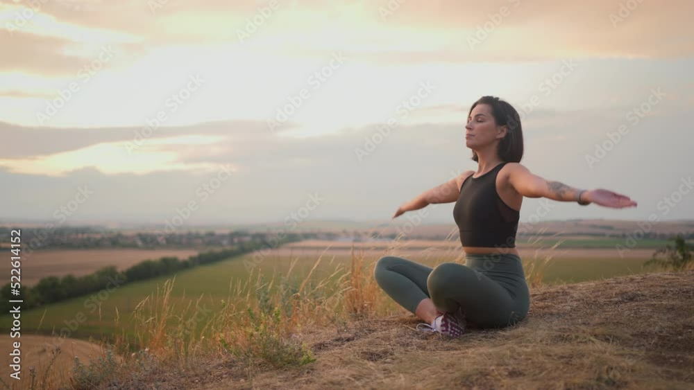 Young beautiful woman doing sport fitness yoga position on a grassy hill during the evening