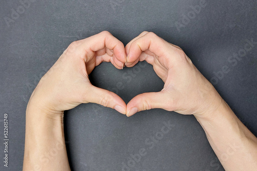 Women s hands are folded in the form of a heart on a gray background. View from above.