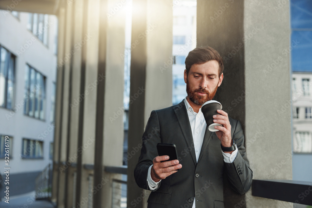 Focused european businessman in suit with phone drinking coffee near office building during break Focused european businessman in suit with phone drinking coffee near office building during break