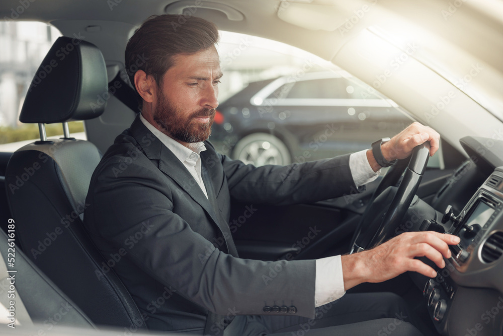 Handsome businessman in grey suit is riding behind steering wheel of car and turns on music Handsome businessman in grey suit is riding behind steering wheel of car and turns on music