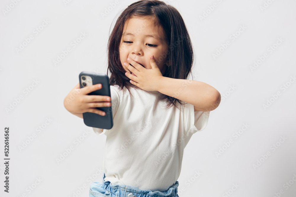 a happy little girl stands with a fashionable smartphone on outstretched arms and looks into her phone covering her mouth with her hand.