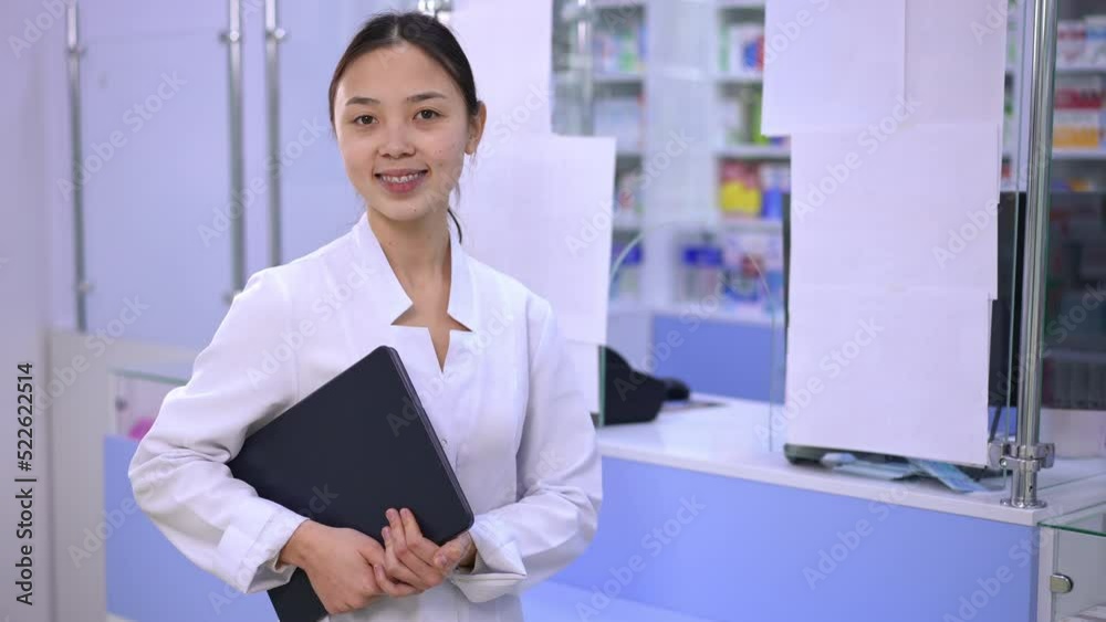 Female Asian pharmacist cashier posing with digital tablet at cash desk in drugstore. Portrait of positive confident beautiful young woman looking at camera smiling standing in pharmacy indoors Female Asian pharmacist cashier posing with digital tablet at cash desk in drugstore. Portrait of positive confident beautiful young woman looking at camera smiling standing in pharmacy indoors