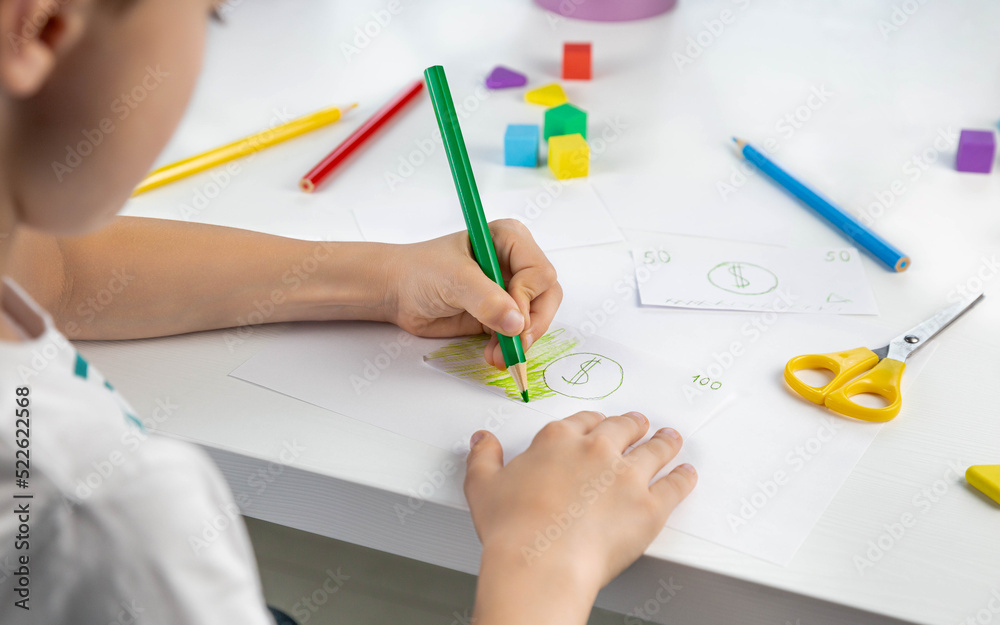 A little boy enthusiastically draws toy money with colored pencils for the game. Family budget. Economy. Money. The kid draws. Economic crisis. Kids play. Selective focus. A little boy enthusiastically draws toy money with colored pencils for the game. Family budget. Economy. Money. The kid draws. Economic crisis. Kids play. Selective focus.