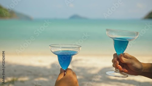 The love couple is holding a glass of blue curacao cocktail, on sea. Close up. Man and woman drink alcohol on sand beach in shadow by azure ocean of a tropical island. Celebrate summer holiday.