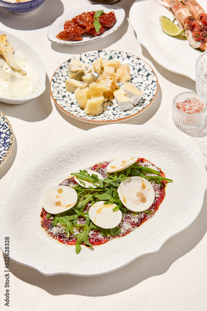 Italian antipasti beef carpaccio with parmesan, ruccola and mushrooms on white table. Carpaccio with raw meat and cheese in italian style with sunny shadows. Italian menu. Aesthetic food composition.