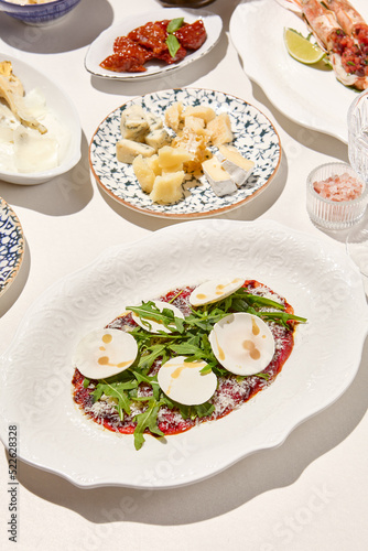 Italian antipasti beef carpaccio with parmesan, ruccola and mushrooms on white table. Carpaccio with raw meat and cheese in italian style with sunny shadows. Italian menu. Aesthetic food composition.