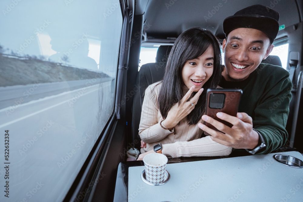 Fototapeta premium suprised couple using smartphone together while traveling by car sitting by the window