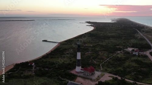 An aerial view of the Fire Island Lighthouse on Long Island, NY. The camera truck left and pan right facing the sunrise and the lighthouse. The light of the lighthouse is on and rotating.