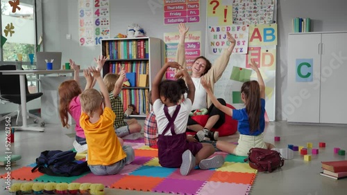 Elementary school teacher sit in class on floor with little pupils and moving hands up