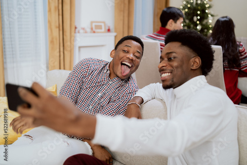 Elegantly dressed man with afro pulls out phone to take selfie with friend, cousin in shirt, brothers smile, silly funny photos in background family eats Christmas Eve dinner
