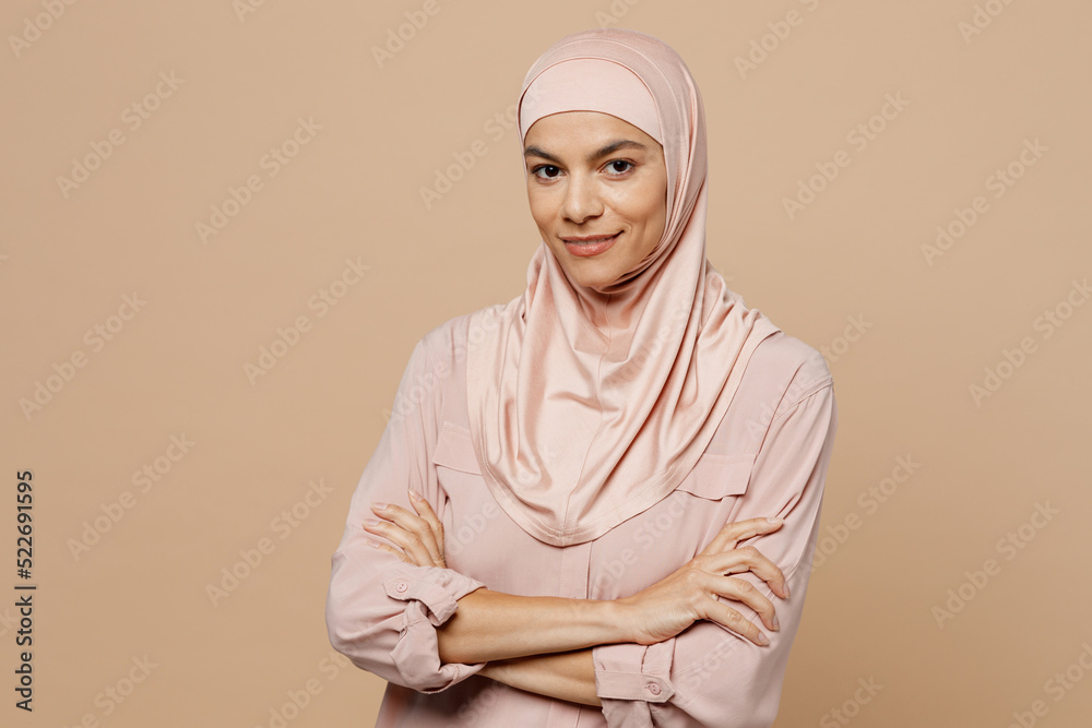 Young smiling arabian asian muslim woman she wear abaya hijab pink clothes hold hands crossed folded isolated on plain pastel light beige background. People uae middle eastern islam religious concept. Young smiling arabian asian muslim woman she wear abaya hijab pink clothes hold hands crossed folded isolated on plain pastel light beige background. People uae middle eastern islam religious concept.