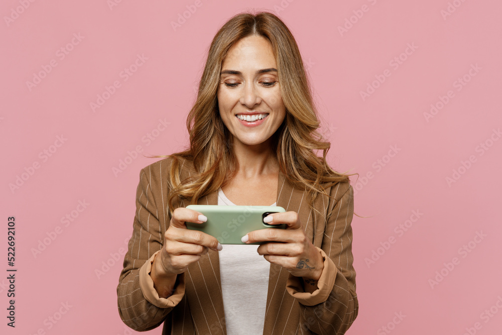 Young employee business woman 30s she wear casual classic jacket using play racing app on mobile cell phone hold gadget smartphone for pc video games isolated on plain pastel light pink background. Young employee business woman 30s she wear casual classic jacket using play racing app on mobile cell phone hold gadget smartphone for pc video games isolated on plain pastel light pink background.