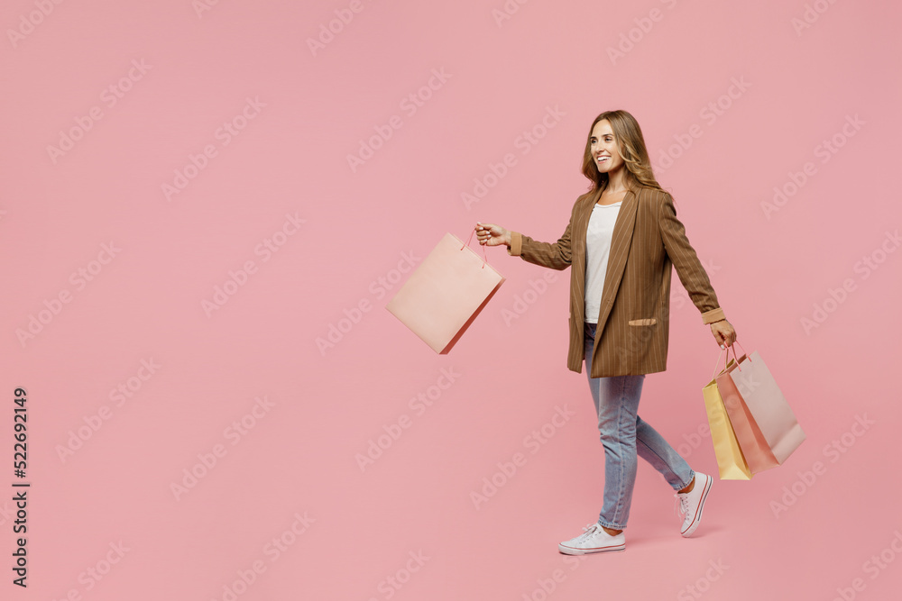 Full body young successful employee business woman 30s she wear casual brown classic jacket hold package bags with purchases after shopping go isolated on plain pastel light pink background studio. Full body young successful employee business woman 30s she wear casual brown classic jacket hold package bags with purchases after shopping go isolated on plain pastel light pink background studio.
