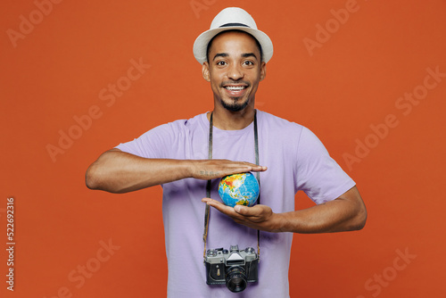 Traveler black man wear purple t-shirt hat hold in palms Earth world globe isolated on plain orange color background. Tourist travel abroad in spare time rest getaway. Air flight trip journey concept. #522692316