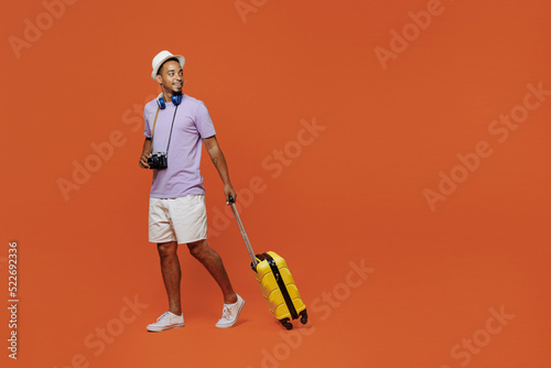 Full size traveler black man wear purple t-shirt hat headphones go with suitcase isolated on plain orange background. Tourist travel abroad in spare time rest getaway. Air flight trip journey concept. #522692336