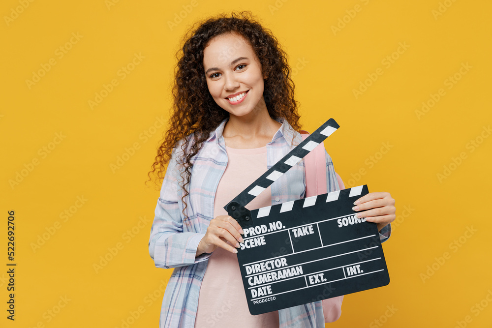Young fun black teen girl student she wear casual clothes backpack bag holding classic black film making clapperboard isolated on plain yellow color background. High school university college concept.