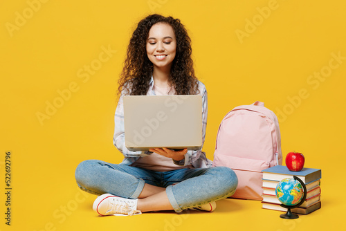 Full body fun young black teen girl student she wear casual clothes backpack bag hold use work on laptop pc computer isolated on plain yellow color background. High school university college concept.