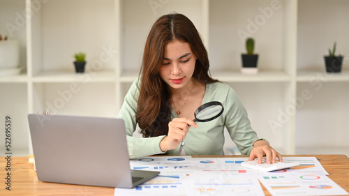 Focused female auditor examining the numerical data on financial document through magnifying glass