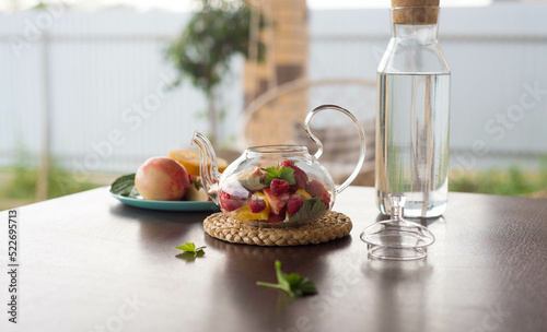A glass teapot with fruits and berries, mint leaves, a bottle of water on the table outside. Summer fruit tea.