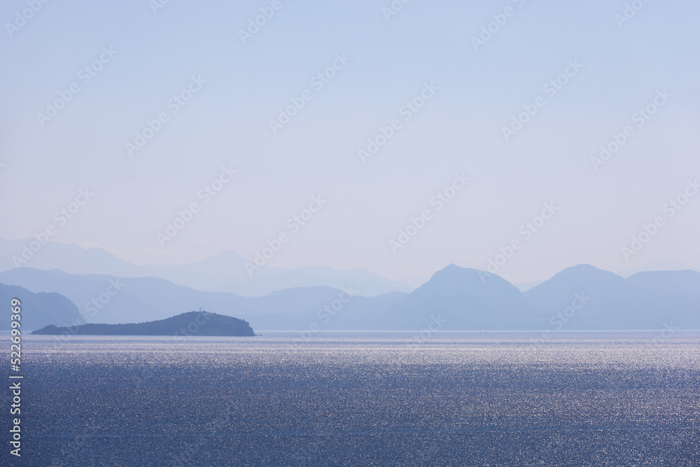 View to blue sea and mountain islands in mist on horizon. Shimmering water surface, background for travelling and vacation