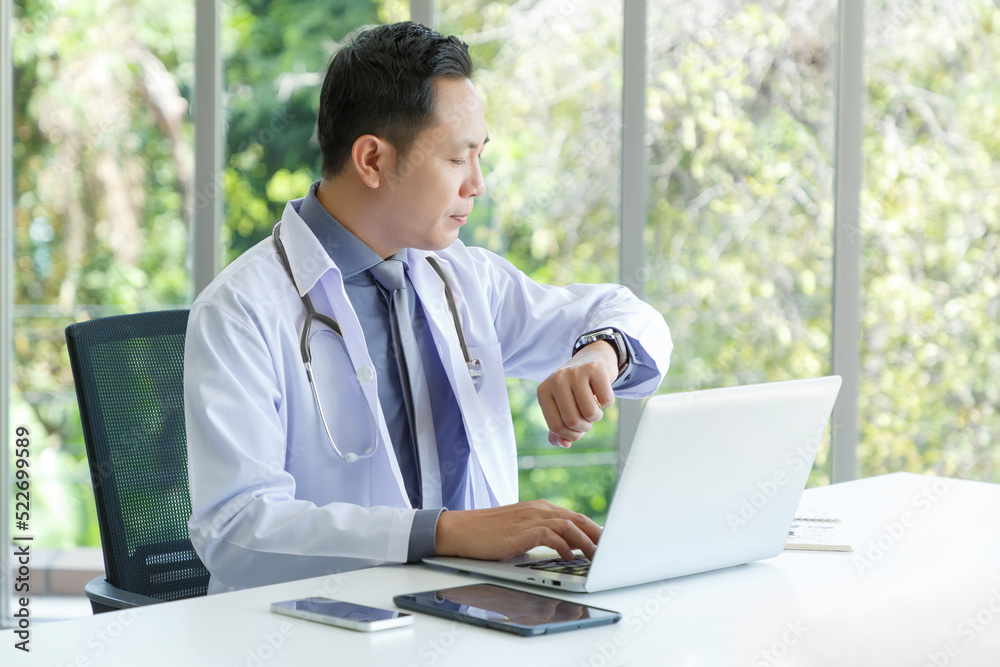 Portrait of asian senior doctor sitting in medical office while using laptop computer and checking time. Portrait of asian senior doctor sitting in medical office while using laptop computer and checking time.