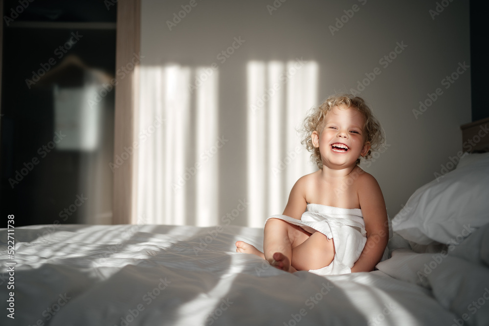 child in a hotel with a smile and an emotion of happiness sits on a bed in a towel with morning rays of light. Vacation child in a hotel with a smile and an emotion of happiness sits on a bed in a towel with morning rays of light. Vacation