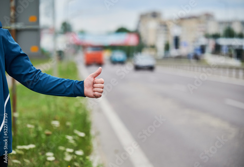 Close up of man's hand hitchhiking by roadside. Male hand showing thumbs up gesture outdoors on blurred background. Hitchhiking, hitching, auto stop concept.