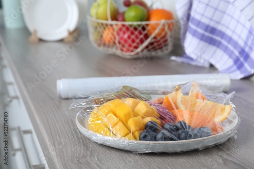 Fototapeta Naklejka Na Ścianę i Meble -  Plate of fresh fruits and berries with plastic food wrap on wooden table indoors