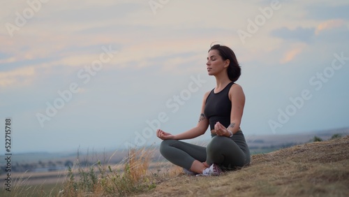 Young beautiful woman doing sport fitness yoga position on a grassy hill during the evening