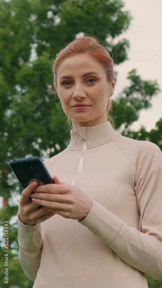 Vertical Screen: Female taking break from jogging and using smartphone, looking into camera and smiles. Woman using mobile phone while doing sports in the park. Vertical Screen: Female taking break from jogging and using smartphone, looking into camera and smiles. Woman using mobile phone while doing sports in the park.