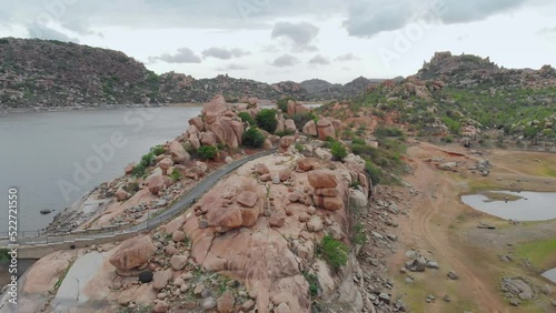 A drone aerial shot of a small curvy road passing through a hilly region with boulders and a river on a cloudy day  photo