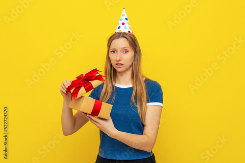 Young shocked woman in a birthday cap and blue t shirt opening gift box and looking impressed with disbelief. Holidays and birthday concept. Colorful studio portrait with yellow background