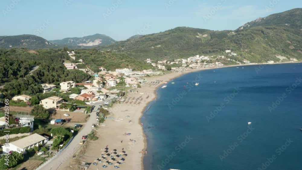 Aerial shot of a Mediterranean beach, right below the green forest covered mountains. Some buildings and beach facilities visible, among sun beds and umbrellas. Aerial shot of a Mediterranean beach, right below the green forest covered mountains. Some buildings and beach facilities visible, among sun beds and umbrellas.