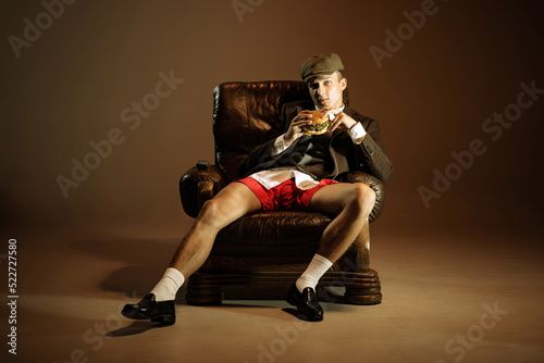 Young man, retro 1920s style english gangster jacket, shorts and cap tasting fast food isolated over dark vintage background. Business, family, art, fashion