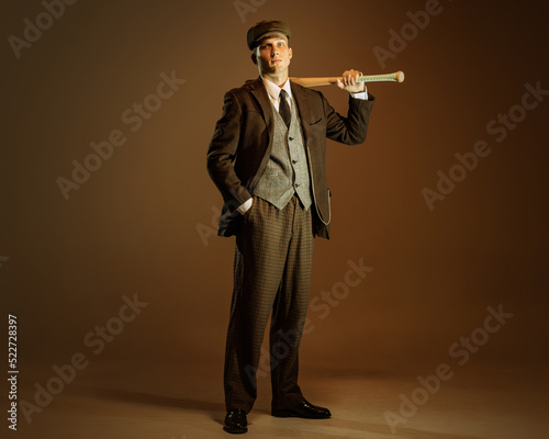 Φωτογραφία Retro style portrait of young man in image of english gangster, businessman wearing suit and cap standing isolated over dark vintage background