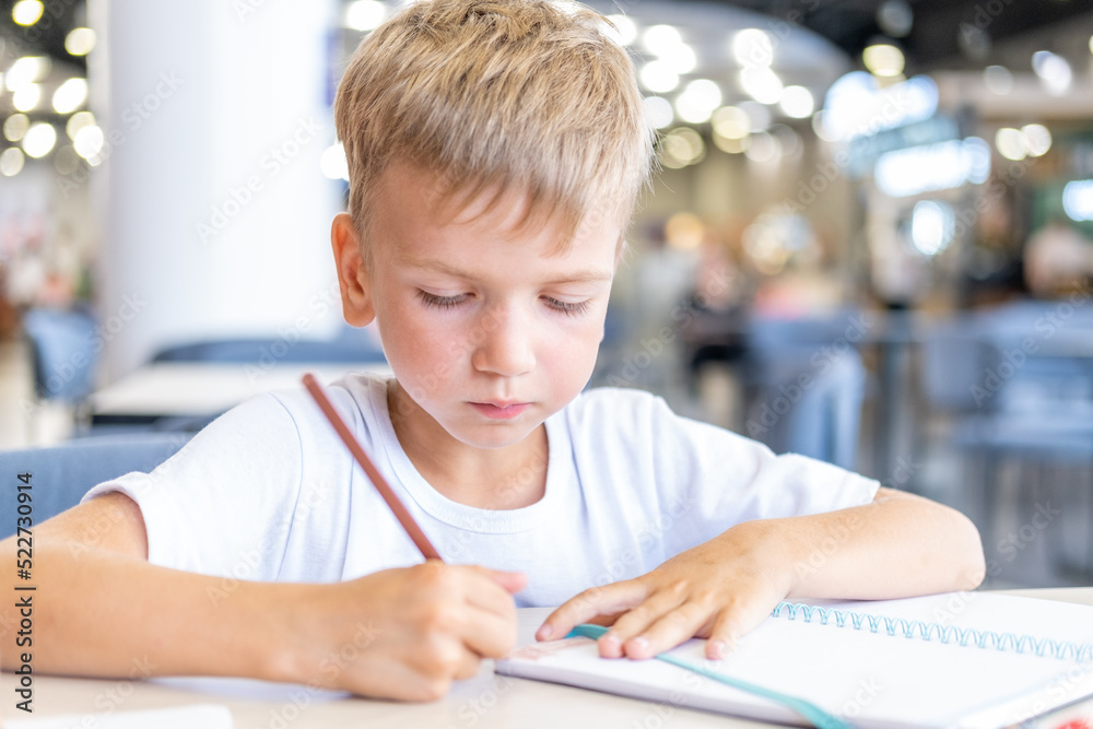 A focused boy doing homework, writing text in a notebook at the table. Cute positive boy writes and smiles. Back to school concept. School and preschool education. A focused boy doing homework, writing text in a notebook at the table. Cute positive boy writes and smiles. Back to school concept. School and preschool education.