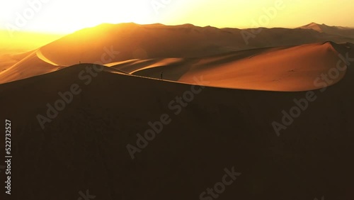 Desert hiker walking, hiking and climbing along a peak of Namibian sand dune. Drone point of view of man enjoying freedom, endless space and sunset or sunrise in remote wild with dry and sandy hills