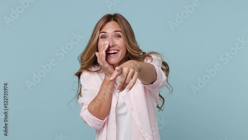 Young excited cheerful woman 30s wear pink shirt look camera laugh smiling watch comedy movie pointing index finger on you isolated on plain pastel light blue cyan background. People lifestyle concept