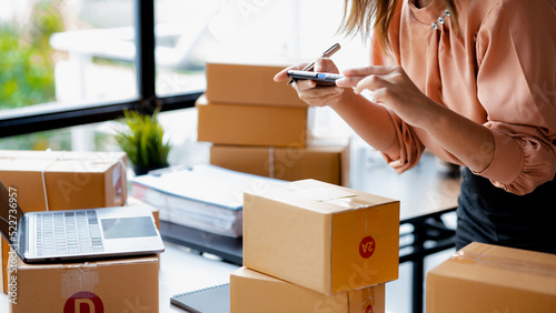Schilderij op canvas A woman using a smartphone to take pictures in front of parcel boxes, parcel boxes for packing goods, delivering goods through private courier companies