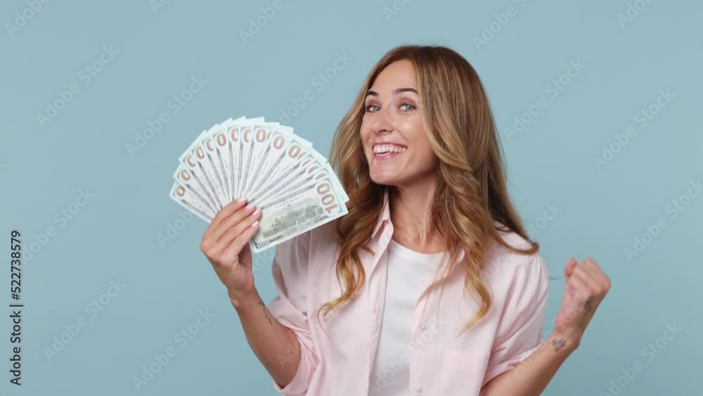Young excited happy woman 30s wear pink shirt hold in hand fan of cash money in dollar banknotes do winner gesture isolated on plain pastel light blue cyan background studio. People lifestyle concept