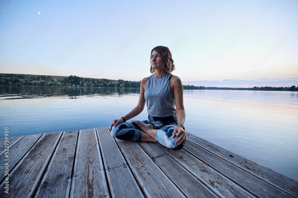 A calm yogi woman in a lotus position is meditating on the dock.