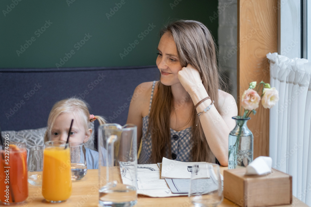 Young mother with daughter sit at table in cafe. Mom is relaxing in restaurant with her small daughter. Young mother with daughter sit at table in cafe. Mom is relaxing in restaurant with her small daughter.