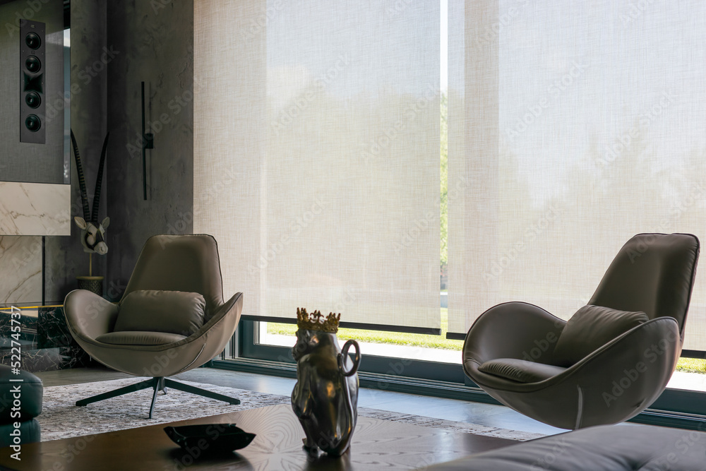 Roller blinds in the interior. Automatic solar shades of large sizes on the  window. Fabric with linen texture. In front of a large window is a chair on  a carpet. Stock Photo