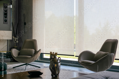 Roller blinds in the interior. Automatic solar shades of large sizes on the window. Fabric with linen texture. In front of a large window is a chair on a carpet. 