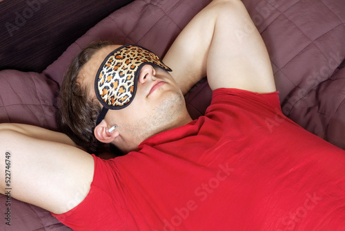 Young handsome man with leopard sleeping mask lying down on the bed, white mobile earphones, red shirt, sleeps or rests. Copy space. photo