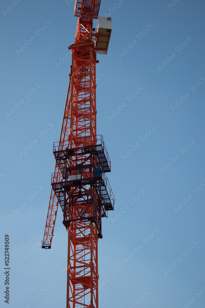 A red orange construction tall crane against a clear blue blue sky in the sunshine of the day in the rays of the sun. Construction site on the background blue sky, panoramic mock-up