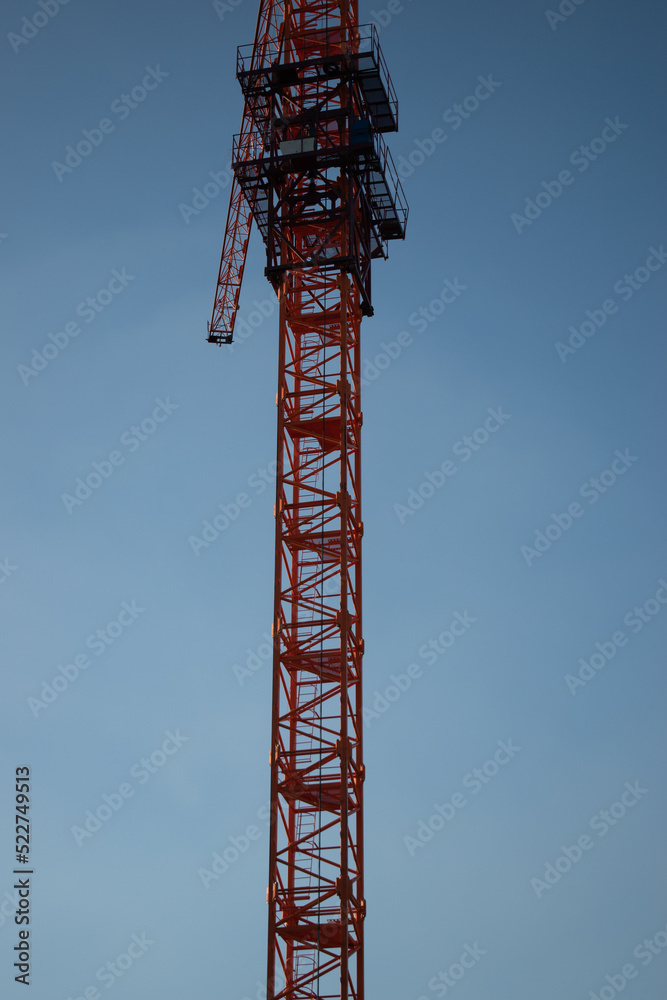 A red orange construction tall crane against a clear blue blue sky in the sunshine of the day in the rays of the sun. Construction site on the background blue sky, panoramic mock-up A red orange construction tall crane against a clear blue blue sky in the sunshine of the day in the rays of the sun. Construction site on the background blue sky, panoramic mock-up