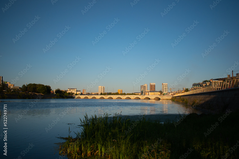 The landscape of the river and bridge, the city in golden sunlight. Grass by the river. Skyscrapers. Clear blue sky. Background, nature. Sunset or sunrise.