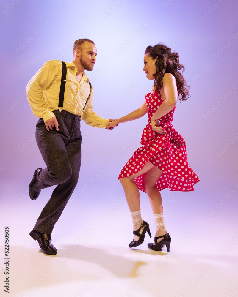 Fototapeta premium Active and emotional couple in colorful retro style costumes dancing incendiary dances isolated on purple background in neon light. Concept of art, culture