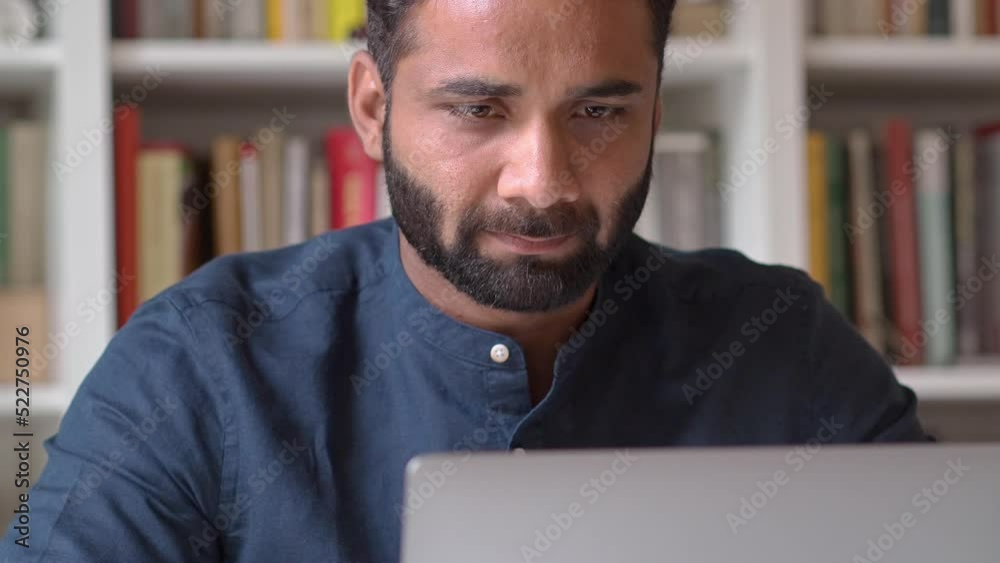 Busy serious indian business man manager remote working or learning online looking at laptop sitting in office at home using software on computer thinking on question analyzing market data technology.